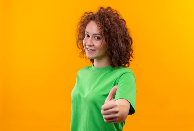 Mujer joven con pelo corto y rizado en camiseta verde sonriendo alegremente mostrando los pulgares para arriba de pie sobre la pared naranja
