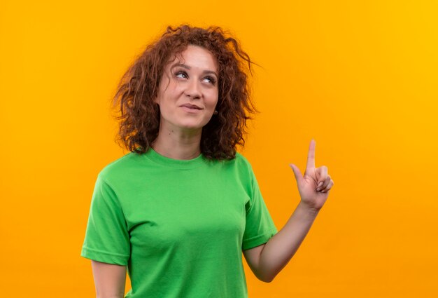 Mujer joven con pelo corto y rizado en camiseta verde sonriendo apuntando hacia arriba con el dedo índice de pie sobre la pared naranja
