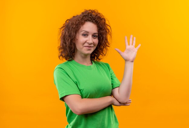 Mujer joven con pelo corto y rizado en camiseta verde sonriendo saludando con la mano de pie sobre la pared naranja