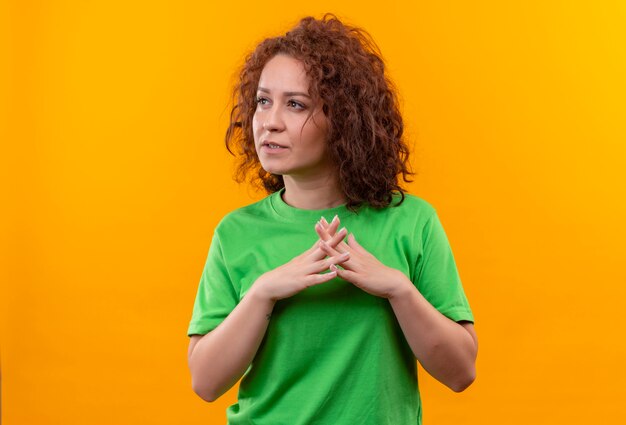 Mujer joven con pelo corto y rizado en camiseta verde sosteniendo las palmas juntas mirando a un lado con expresión pansive de pie