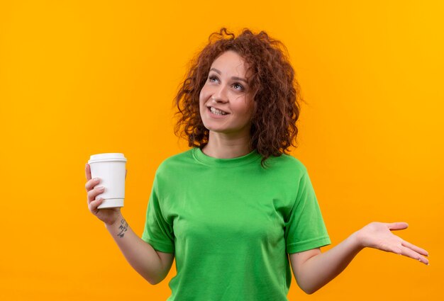 Mujer joven con pelo corto y rizado en camiseta verde sosteniendo la taza de café mirando hacia arriba sonriendo extendiendo el brazo hacia el lado de pie sobre la pared naranja