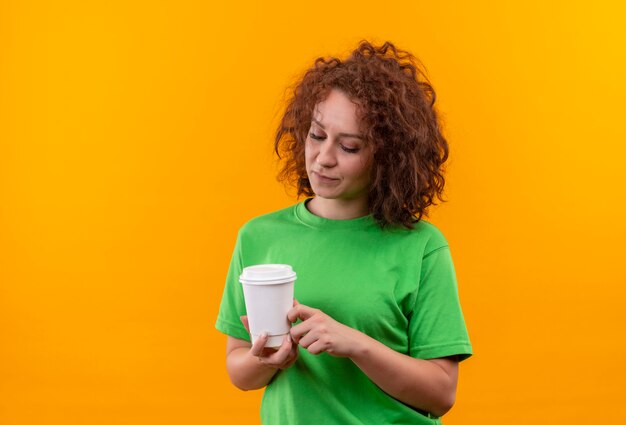 Mujer joven con pelo corto y rizado en camiseta verde sosteniendo la taza de café mirándola con expresión triste en la cara de pie sobre la pared naranja