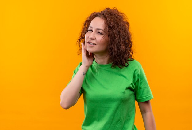 Mujer joven con pelo corto y rizado en camiseta verde tocando su mejilla sintiendo dolor de pie sobre la pared naranja