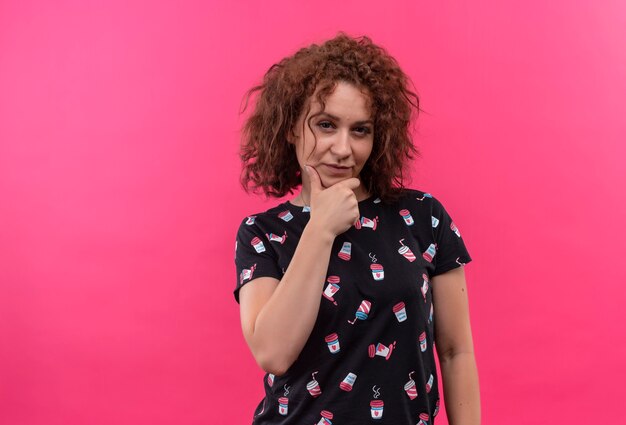 Mujer joven con pelo corto y rizado mirando con expresión pensativa en la cara pensando de pie sobre la pared rosa