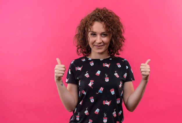 Mujer joven con pelo corto y rizado sonriendo mostrando los pulgares para arriba con ambas manos de pie sobre la pared rosa