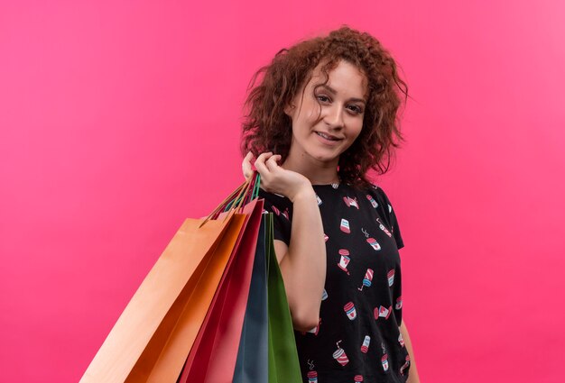 Mujer joven con pelo corto y rizado sosteniendo bolsas de papel sonriendo alegremente feliz y positivo parado sobre pared rosa