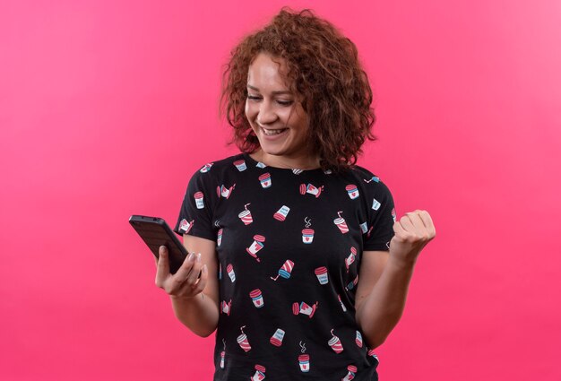Mujer joven con pelo corto y rizado sosteniendo smartphone mirando la pantalla feliz y emocionado puño de pie sobre la pared rosa
