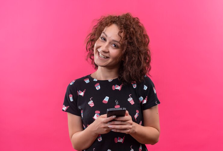 Foto gratuito mujer joven con pelo corto y rizado sosteniendo smartphone sonriendo alegremente parado sobre pared rosa