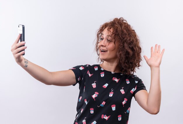 Mujer joven con pelo corto y rizado tomando selfie con su teléfono inteligente sonriendo a la cámara saludando con la mano de pie sobre la pared blanca