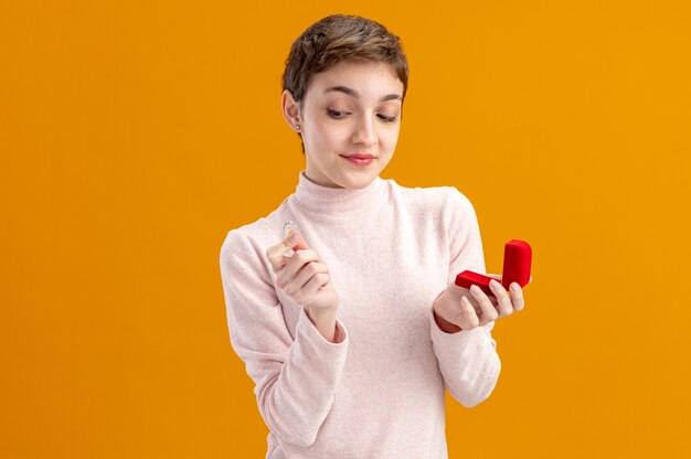 Mujer joven con pelo corto sosteniendo el cuadro rojo y el anillo de compromiso mirándolo con una sonrisa en la cara el concepto del día de San Valentín de pie sobre la pared naranja