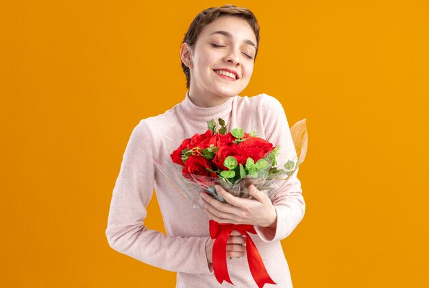 Mujer joven con pelo corto sosteniendo un ramo de rosas rojas feliz y positivo con los ojos cerrados sonriendo concepto del día de San Valentín de pie sobre la pared naranja