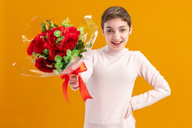 Mujer joven con pelo corto sosteniendo un ramo de rosas rojas mirando a la cámara feliz y positivo sonriendo ampliamente concepto del día de San Valentín de pie sobre la pared naranja