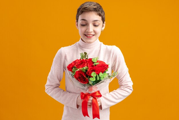 Mujer joven con pelo corto sosteniendo un ramo de rosas rojas mirando rosas con una sonrisa en la cara feliz día de San Valentín concepto de pie sobre la pared naranja