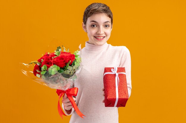 Mujer joven con el pelo corto sosteniendo un ramo de rosas rojas y un presente mirando a la cámara feliz y positivo sonriendo alegremente el concepto del día de San Valentín de pie sobre la pared naranja