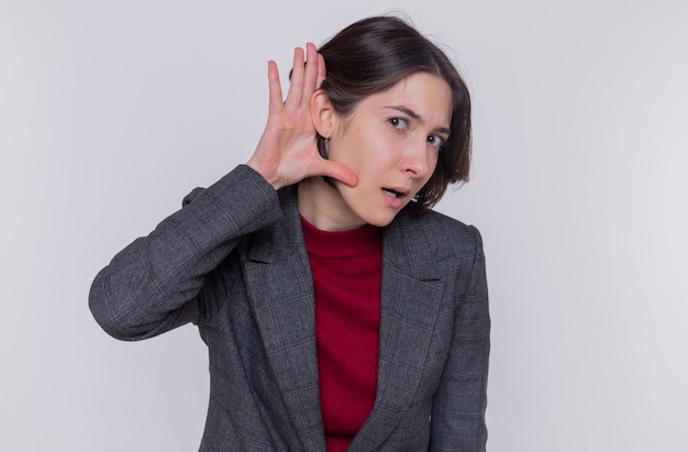 Mujer joven con pelo corto vistiendo chaqueta gris mirando intrigado sosteniendo la mano sobre la oreja tratando de escuchar de pie sobre la pared blanca