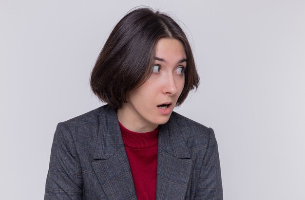 Mujer joven con pelo corto vistiendo chaqueta gris mirando a un lado asombrado y sorprendido de pie sobre la pared blanca