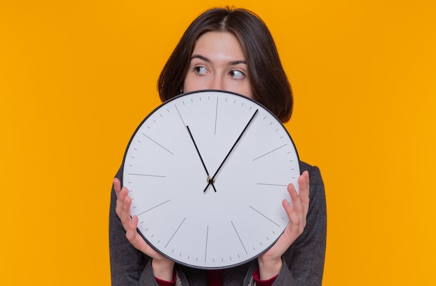 Foto gratuita mujer joven con pelo corto vistiendo chaqueta gris sosteniendo el reloj de pared mirando a un lado preocupado de pie sobre la pared naranja