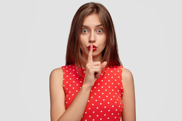 Una mujer joven de pelo largo de aspecto agradable usa lápiz labial rojo, hace un gesto de silencio, pide que no le cuentes a nadie un secreto