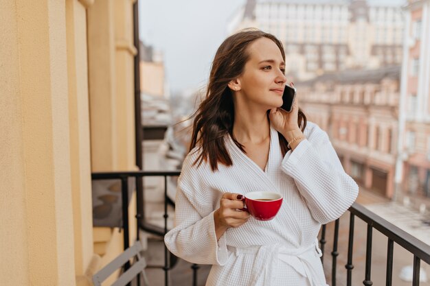 Mujer joven de pelo largo disfrutando de la vista de la ciudad en el balcón. Chica en bata de baño bebe café y habla por teléfono.