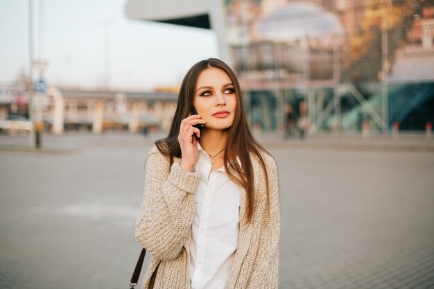 La mujer joven con el pelo largo habla en el teléfono que camina afuera en la tarde