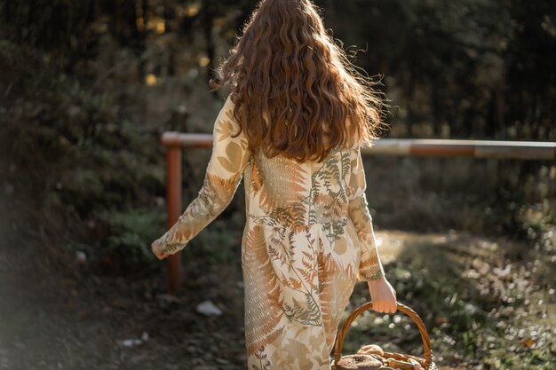 Mujer joven con el pelo largo y rojo en un vestido de lino reuniendo setas en el bosque