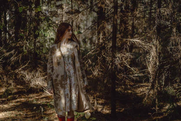 Mujer joven con el pelo largo y rojo en un vestido de lino reuniendo setas en el bosque