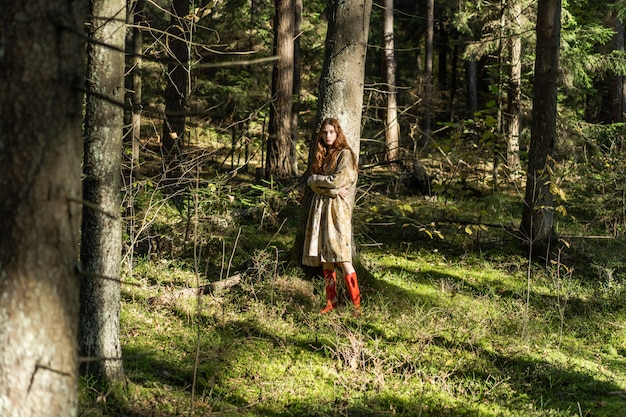 Mujer joven con el pelo largo y rojo en un vestido de lino reuniendo setas en el bosque