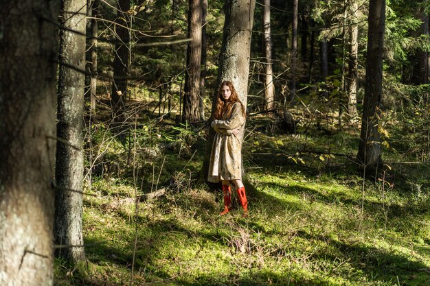 Mujer joven con el pelo largo y rojo en un vestido de lino reuniendo setas en el bosque