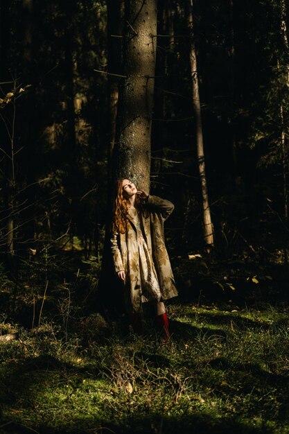 Mujer joven con el pelo largo y rojo en un vestido de lino reuniendo setas en el bosque