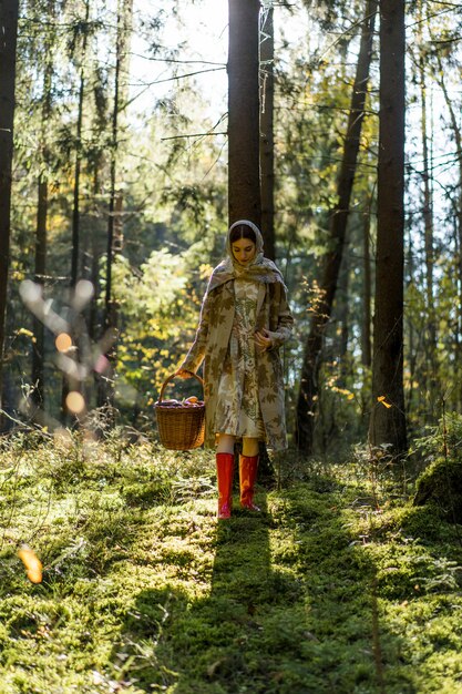 Mujer joven con el pelo largo y rojo en un vestido de lino reuniendo setas en el bosque