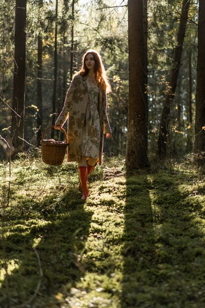 Mujer joven con el pelo largo y rojo en un vestido de lino reuniendo setas en el bosque