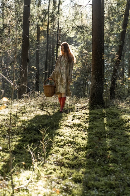 Mujer joven con el pelo largo y rojo en un vestido de lino reuniendo setas en el bosque