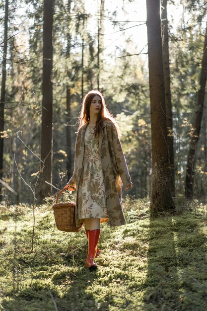 Mujer joven con el pelo largo y rojo en un vestido de lino reuniendo setas en el bosque