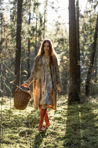 Mujer joven con el pelo largo y rojo en un vestido de lino reuniendo setas en el bosque