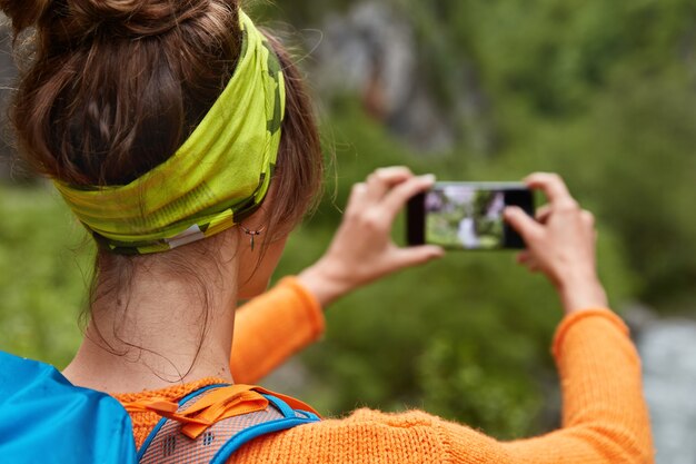 Mujer joven de pelo oscuro retrocede, lleva diadema verde, lleva mochila, hace fotos en el dispositivo smartphone