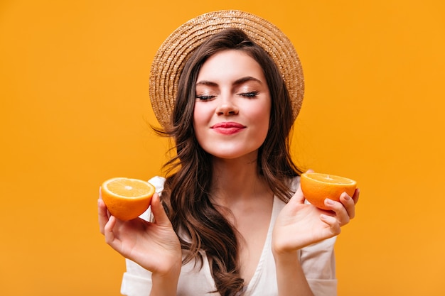 Mujer joven de pelo oscuro rizado en navegante sostiene naranjas y posa con los ojos cerrados.
