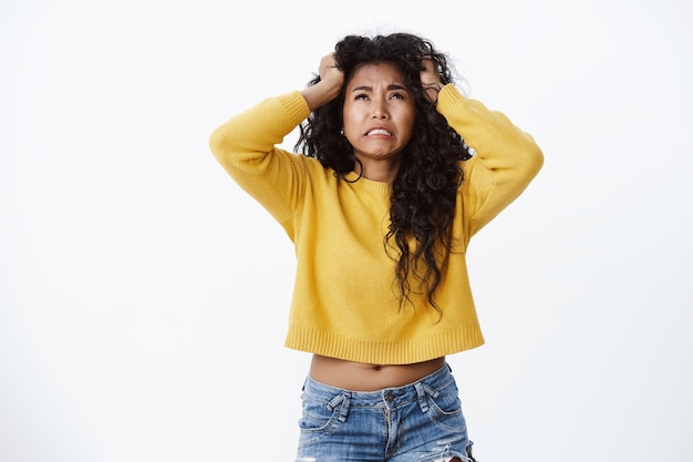 Foto gratuita mujer joven de pelo rizado devastada y preocupada en suéter amarillo recortado en pánico, sintiéndose incómoda y preocupada, mirar hacia arriba deprimida, tomarse de las manos en la cabeza molesta, pared blanca