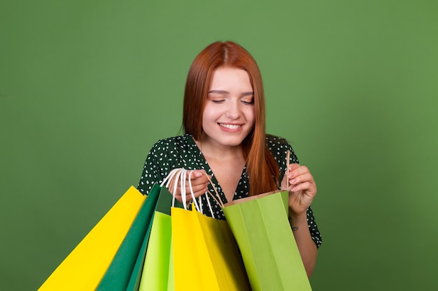 Mujer joven de pelo rojo en la pared verde con bolsas de compras feliz alegre emocionado