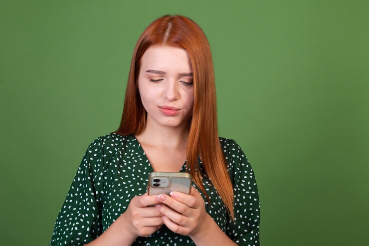 Foto gratuito mujer joven de pelo rojo en la pared verde con mensajes de texto de teléfono móvil charlando con una sonrisa en la cara, feliz positivo