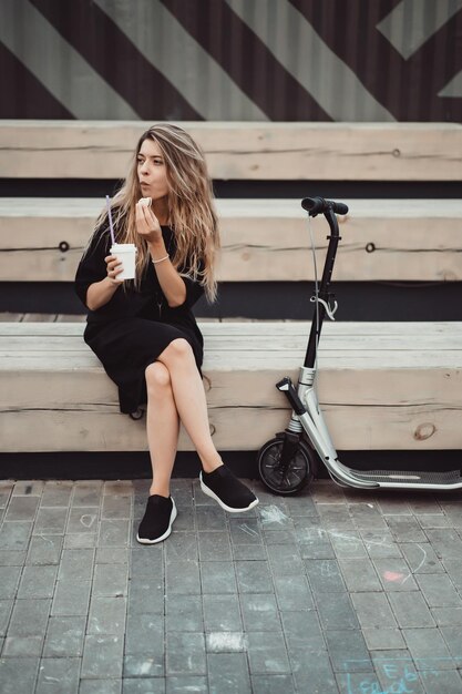 Mujer joven con pelos largos en scooter eléctrico. La niña en el scooter eléctrico bebe café.