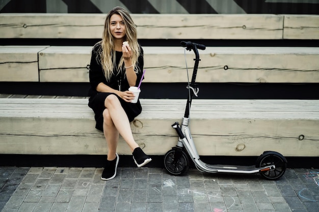 Mujer joven con pelos largos en scooter eléctrico. La niña en el scooter eléctrico bebe café.