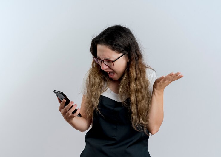 Foto gratuito mujer joven peluquera profesional en delantal mirando la pantalla de su teléfono inteligente volviéndose loca gritando con expresión agresiva de pie sobre la pared blanca