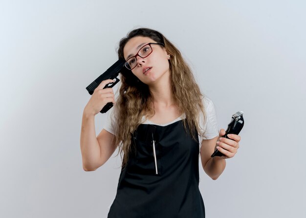 Mujer joven peluquera profesional en delantal sosteniendo recortadora y pistola cerca de su sien mirando a un lado cansado y aburrido de pie sobre la pared blanca