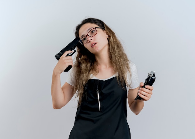Mujer joven peluquera profesional en delantal sosteniendo recortadora y pistola cerca de su sien mirando a un lado cansado y aburrido de pie sobre la pared blanca