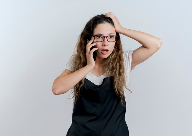 Mujer joven peluquero profesional en delantal hablando por teléfono móvil confundido de pie sobre la pared blanca