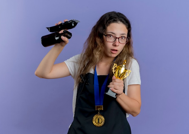 Mujer joven peluquero profesional en delantal con medalla de oro alrededor del cuello sosteniendo recortadora y trofeo mirando al frente preocupado de pie sobre la pared azul