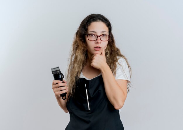 Mujer joven peluquero profesional en delantal sosteniendo recortadora mirando al frente con la mano en la barbilla pensando de pie sobre la pared blanca