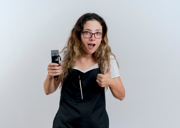 Mujer joven peluquero profesional en delantal sosteniendo recortadora mirando al frente sonriendo mostrando los pulgares para arriba de pie sobre la pared blanca