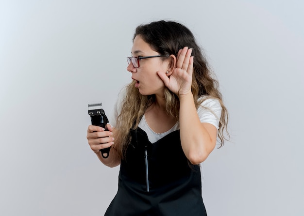 Mujer joven peluquero profesional en delantal sosteniendo recortadora sosteniendo la mano cerca de su oreja tratando de escuchar chismes de pie sobre la pared blanca