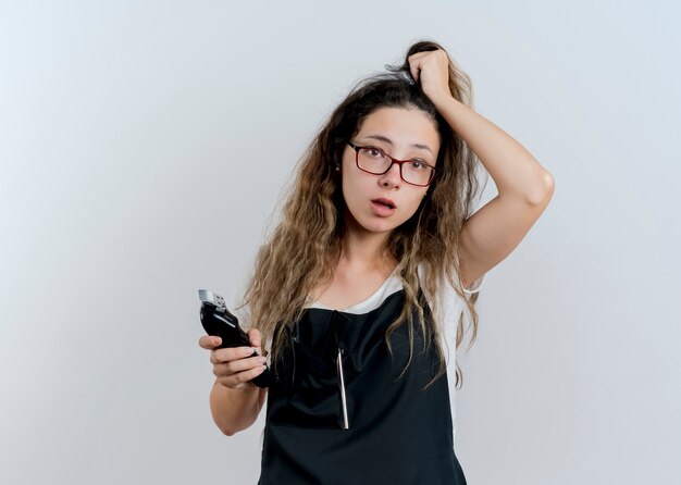 Mujer joven peluquero profesional en delantal sosteniendo recortadora tocando su cabello mirando confundido de pie sobre la pared blanca